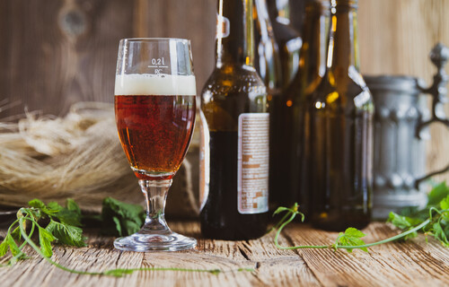 Nicht ganz volles kleines Bierglas vor dunklen Bierflaschen auf Holztisch | © Land schafft Leben